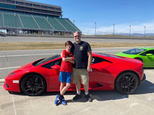 A man and a child pose together beside a red Lamborghini at a racetrack on a sunny day.