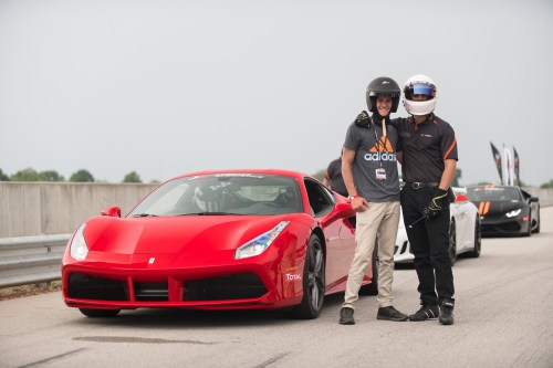 Two people pose beside a red sports car on a racetrack, one wearing a helmet and the other in casual attire.