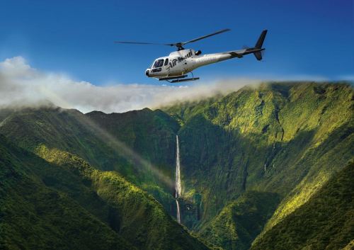 A helicopter flies over lush green mountains with a waterfall cascading down in the background.
