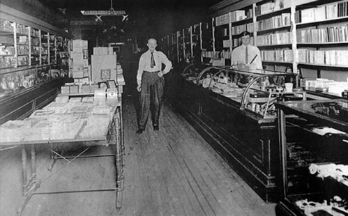 Black and white photo of a vintage store interior with two men standing among shelves and display cases.