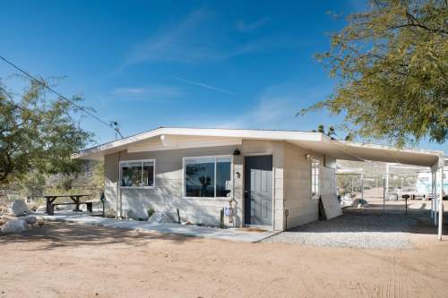 A small, modern house with a gravel driveway, surrounded by desert landscape and trees under a clear blue sky.