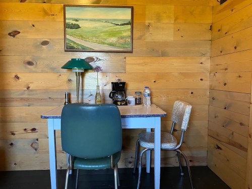 A cozy room with wooden walls, featuring a small table, two chairs, a coffee maker, and a landscape painting.