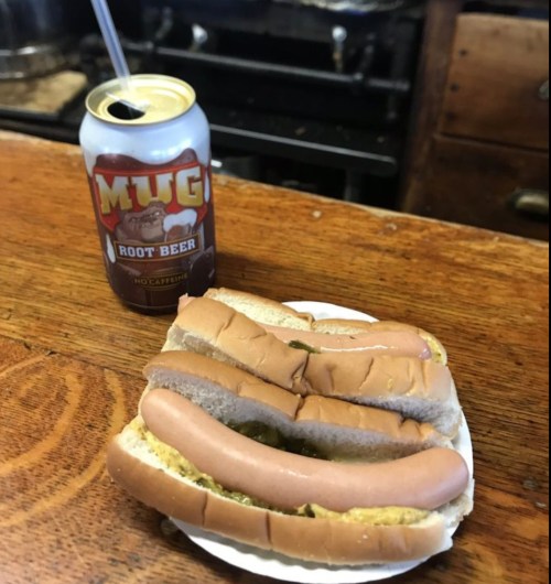 A plate with three hot dogs topped with mustard and pickles, next to a can of Mug Root Beer on a wooden surface.