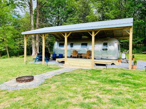 A shiny Airstream trailer under a wooden shelter, surrounded by greenery and a stone path leading to a fire pit.