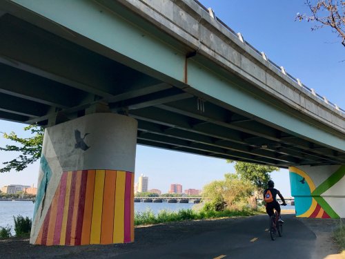 A cyclist rides under a colorful bridge, with murals on the pillars and a river visible in the background.