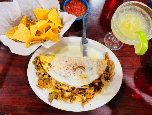 A plate with a large cheesy omelette filled with mushrooms and chicken, served with tortilla chips, salsa, and a margarita.