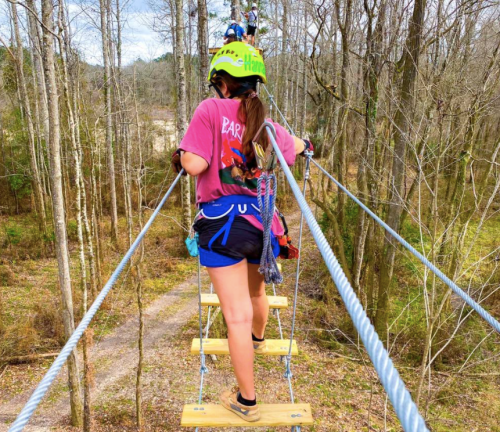 A person in a helmet walks across a zip line platform in a forested area, surrounded by trees and a path below.