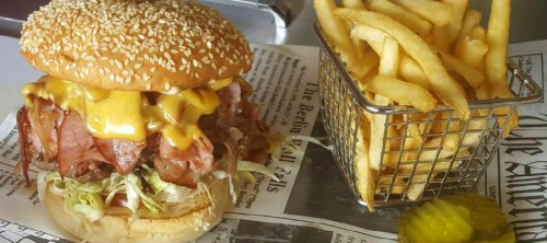A large sesame seed burger stacked with meat, cheese, and lettuce, served with a side of crispy fries in a metal basket.