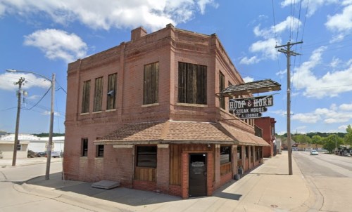 Historic brick building with boarded windows, featuring a sign for "Hoof & Horn Steak House and Lodge" under a blue sky.