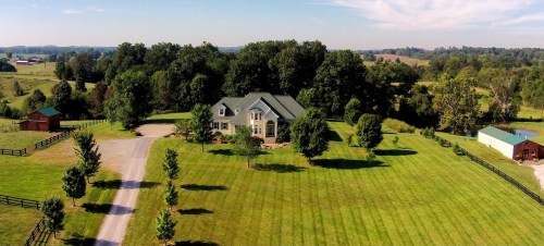 Aerial view of a large house surrounded by green trees and manicured lawns, with a gravel driveway leading up to it.