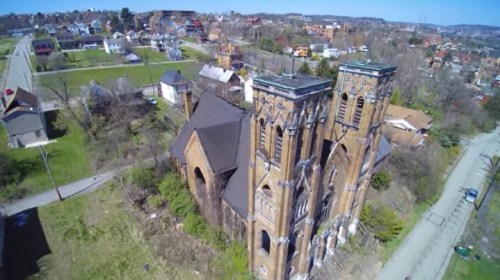 Aerial view of an abandoned church surrounded by overgrown land and residential buildings in a rural area.