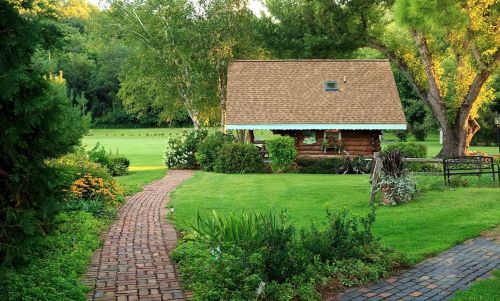 A charming log cabin surrounded by lush greenery and a brick pathway leading through a vibrant garden.