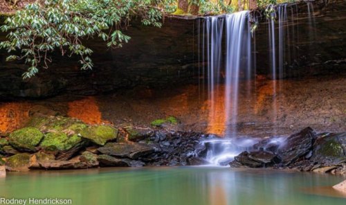 A serene waterfall cascades over rocks into a tranquil pool, surrounded by lush greenery and earthy tones.