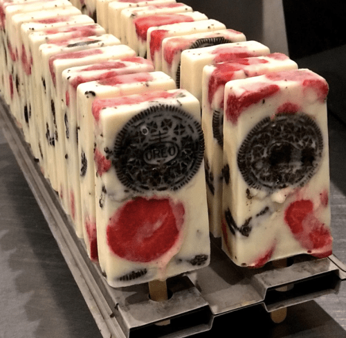A row of colorful ice pops with layers of strawberries and Oreo pieces, displayed on a metal tray.