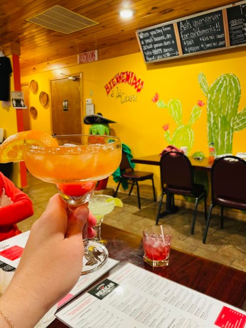 A hand holds a colorful cocktail in a restaurant with a vibrant yellow wall and cactus decor in the background.