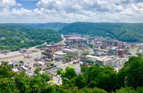 A panoramic view of a town surrounded by green hills, featuring buildings, roads, and a river in the distance.
