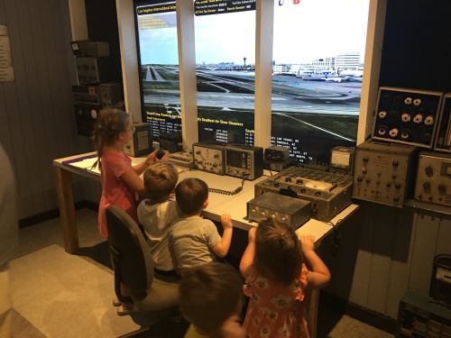 Children interact with a flight simulation setup featuring multiple screens and control equipment in a museum setting.
