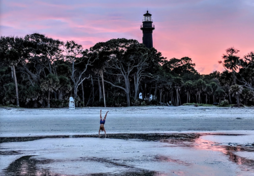 A person doing a handstand on the beach at sunset, with a lighthouse and trees in the background.