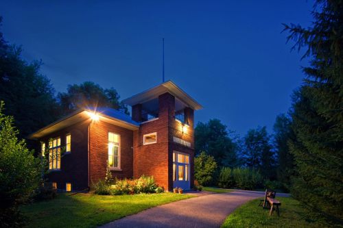 A brick house illuminated at dusk, surrounded by greenery and a pathway leading to the entrance.