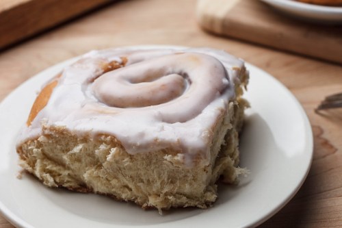 A close-up of a frosted cinnamon roll on a white plate, with a soft, fluffy texture and a glossy icing.