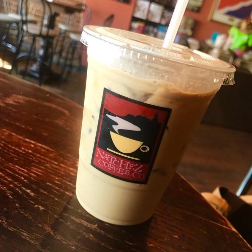 Iced coffee in a clear cup with a logo from Natchez Coffee Co., placed on a wooden table in a café setting.