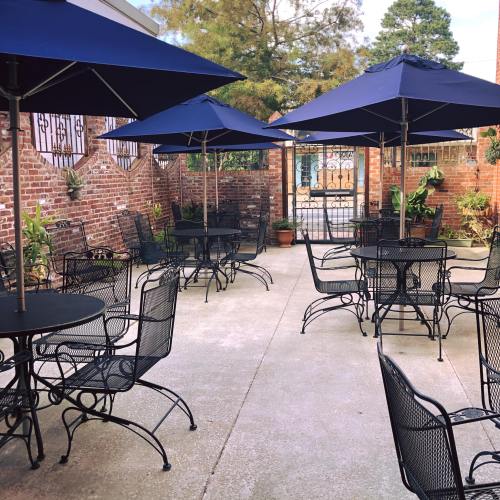 Outdoor patio with black metal tables and chairs, shaded by blue umbrellas, surrounded by brick walls and greenery.