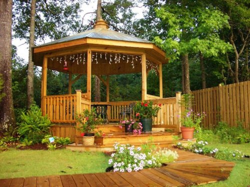 A wooden gazebo surrounded by greenery and flowers, featuring a stone pathway and decorative lights.