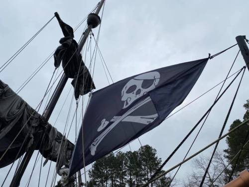 A pirate flag with a skull and crossbones flies from a ship's mast against a cloudy sky.