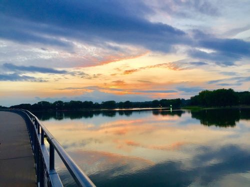 A serene lake at sunset, with colorful clouds reflecting on the water and a railing along the shore.