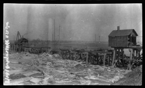 Black and white photo of a wooden pier with a small house on stilts, surrounded by debris and a flat landscape.