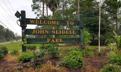 Welcome sign for John Slidell Park, surrounded by greenery and flowers, with a road visible in the background.