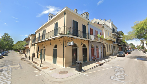 A corner building with a balcony, surrounded by trees and a clear blue sky, on a quiet street in a historic area.