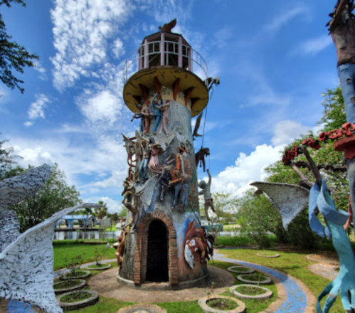 Colorful, whimsical lighthouse sculpture surrounded by greenery and blue sky, featuring various artistic figures and designs.