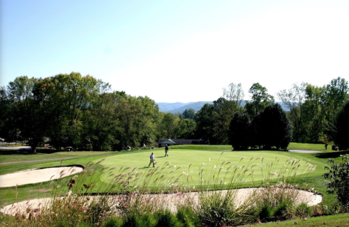 A sunny golf course with two golfers on the green, surrounded by trees and a sand trap in the foreground.