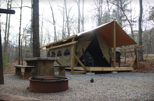 A canvas tent on a wooden platform surrounded by trees, with a picnic table and fire pit nearby.