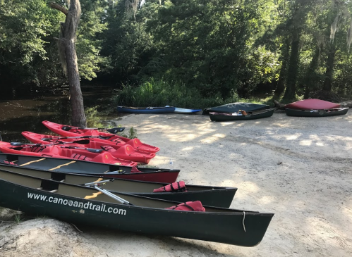 Canoes lined up on a sandy riverbank, with lush greenery and calm water in the background.