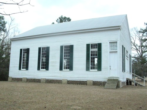 A white wooden building with green shutters and large windows, set on a grassy area with trees in the background.