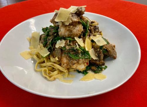 A plate of pasta topped with shrimp, greens, and shaved cheese, served on a white dish against a red background.