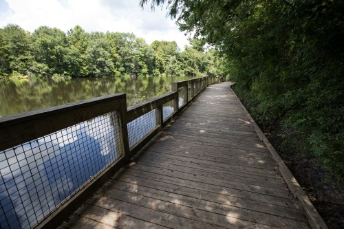 A wooden walkway along a calm lake, surrounded by lush greenery and trees under a partly cloudy sky.