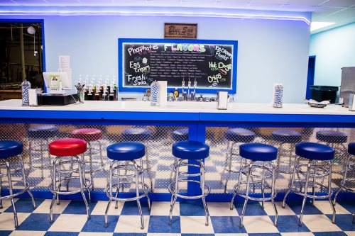A retro diner counter with colorful stools and a chalkboard menu displaying flavors and offerings.