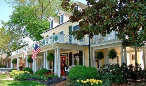 Charming two-story house with a porch, American flag, and colorful flower beds under a clear blue sky.