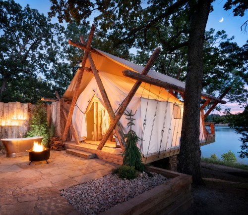 A cozy glamping tent by a lake, featuring a fire pit and surrounded by trees at dusk with a crescent moon in the sky.