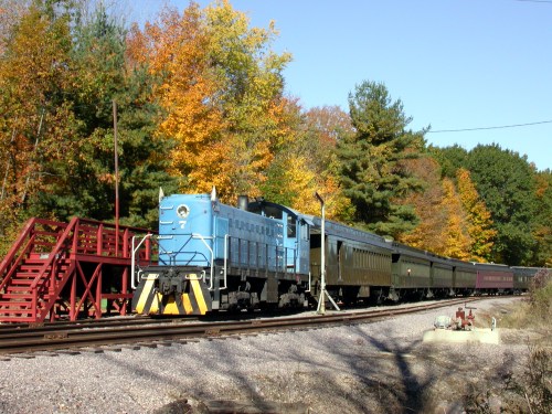 A blue locomotive pulls a vintage train through a colorful autumn landscape with trees in vibrant fall foliage.