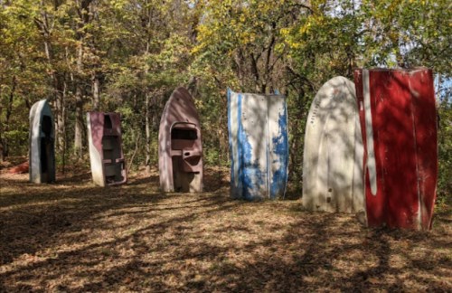 A row of colorful, weathered boat hulls standing upright in a wooded area with fallen leaves on the ground.