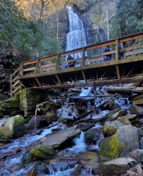 A wooden bridge spans a rocky stream, with a waterfall cascading in the background surrounded by lush greenery.