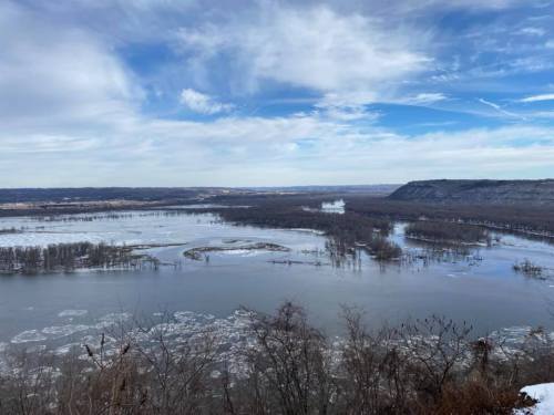 A scenic view of a river with ice patches, surrounded by trees and hills under a cloudy blue sky.