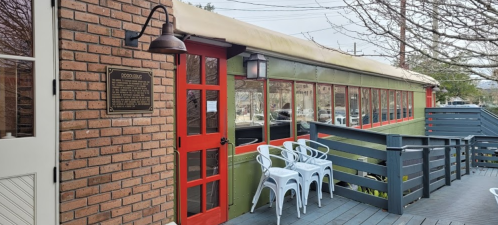A colorful building with large windows, a red door, and a plaque, surrounded by a wooden deck and outdoor seating.
