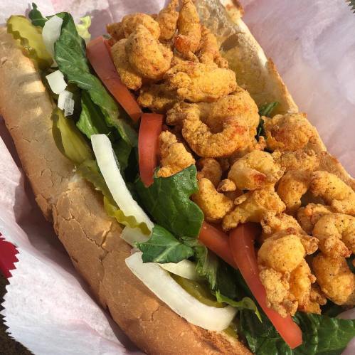 A sandwich filled with crispy fried shrimp, lettuce, tomato, and onions on a hoagie roll, served in a paper wrapper.