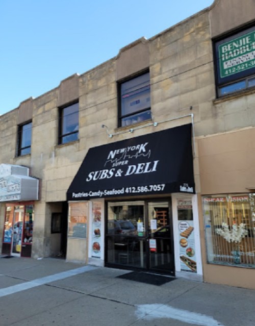 A storefront with a black awning reading "New York Subs & Deli," featuring a menu of pastries, candy, and seafood.