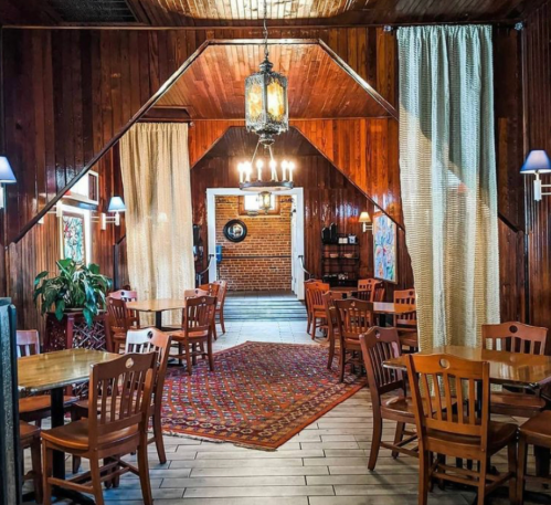 Cozy restaurant interior with wooden walls, tables, and a patterned rug, featuring warm lighting and decorative curtains.